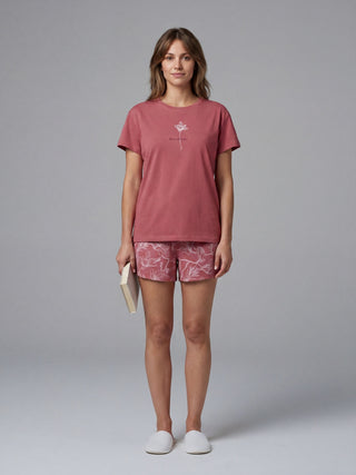 A woman in a pink t-shirt with a floral graphic and pink floral printed pajama shorts stands holding a book, wearing white slippers.