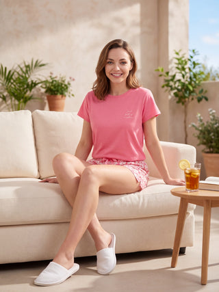 A woman in a pink t-shirt with a "Soul full of sunshine" graphic and matching pink floral print shorts sits on a cream couch, wearing white slippers.