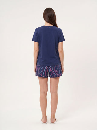 A woman from behind wearing a navy blue t-shirt and navy blue shorts with a colorful abstract stripe print, white slippers.