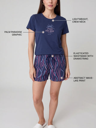 A woman stands in a navy blue pajama set with an abstract wave-like print on the shorts and a "Palm Paradise" graphic on the t-shirt.