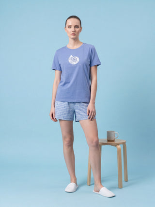 Women lounging on a bed in blue pajama shorts and a periwinkle t-shirt with a white seashell print.