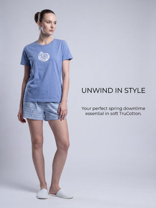 Woman in a light blue short-sleeve t-shirt with a seashell print and matching striped shorts, standing and looking to the side, with white slippers.