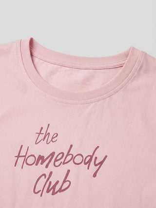 A close-up shot of a woman's pink t-shirt, featuring a delicate cursive print that reads "the Homebody Club."