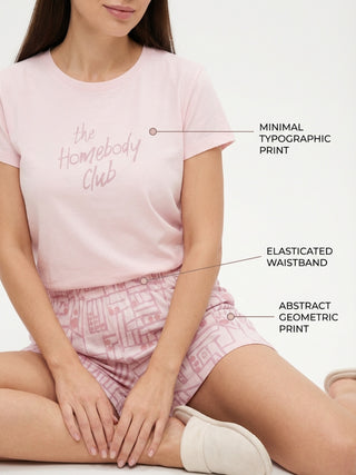 Women, seated cross-legged, wearing a light pink t-shirt with a "Homebody Club" print and matching patterned shorts, featuring an elasticated waistband and abstract geometric print.