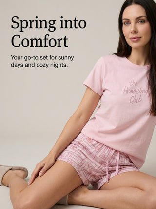 A woman poses in pink loungewear, a t-shirt with "the Homebody Club" print and patterned shorts.