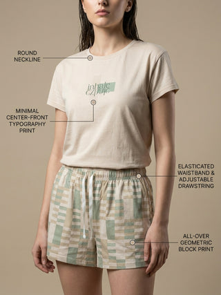 A woman wearing a beige t-shirt with a minimalist center-front typography print and light green and beige geometric block print shorts with an elasticated waistband and adjustable drawstring.