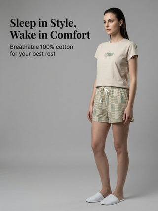 A woman in a cream-colored t-shirt with a green graphic and patterned green and tan shorts poses with white slippers.