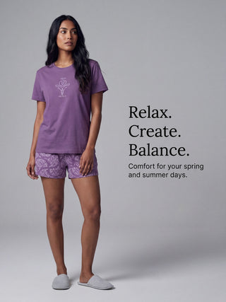 A woman in a purple t-shirt with a white graphic print and matching purple shorts with a white damask print stands comfortably.