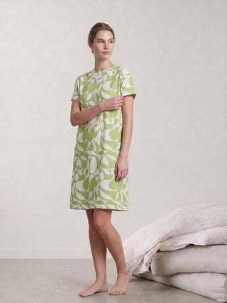 A woman in a light green and white patterned short-sleeve sleep dress sits on a beige couch, wearing white slippers.