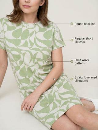 A woman in a light green and white patterned short-sleeve sleep dress sits on a beige couch, wearing white slippers.