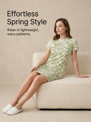 Woman in a green and white wavy print t-shirt dress and white slippers, sitting on a cream sofa.