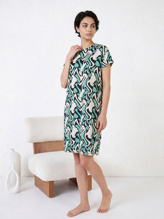 A woman in a green and cream abstract print midi dress and white slippers sits on a beige couch, looking to her left.