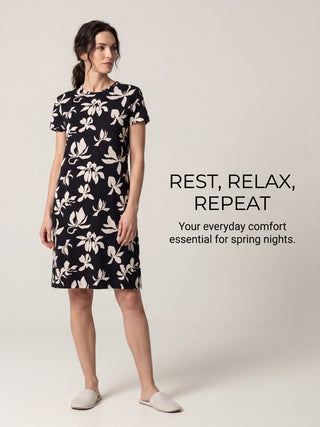 A woman in a black short-sleeved nightgown with a white floral print stands with her left leg forward, posing for a sleepwear advertisement.