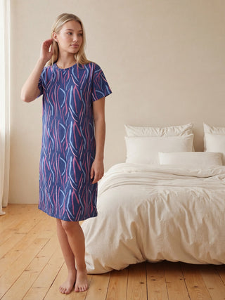 A woman in a navy blue nightgown with a pink and purple wavy print, wearing blue slippers, stands in a relaxed pose.
