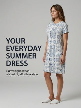 A woman poses in a white short-sleeved dress with a blue floral print and white slides.