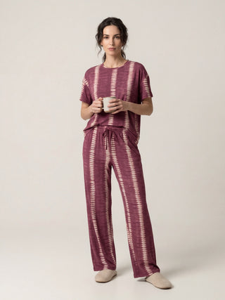 Woman standing, holding a mug, wearing a burgundy tie-dye pajama set and beige slippers.