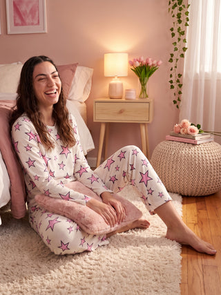 A woman in white pajamas with pink stars laughs while sitting on a fluffy rug.