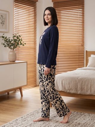 Women in a navy blue long-sleeved sleep top with a light beige and navy blue floral print pajama set, posing side-profile on a rug next to a bed.