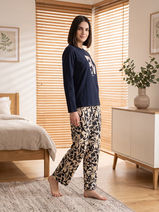 A woman stands in a bedroom wearing a navy blue long-sleeved pajama top with a graphic print and beige pajama pants with a dark floral print.