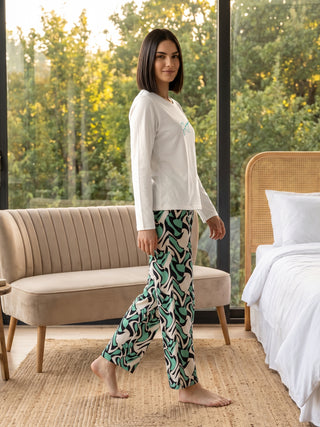 Woman in a white long-sleeved top with a blue graphic and green, black, and white patterned pajama pants, posing with her legs spread.