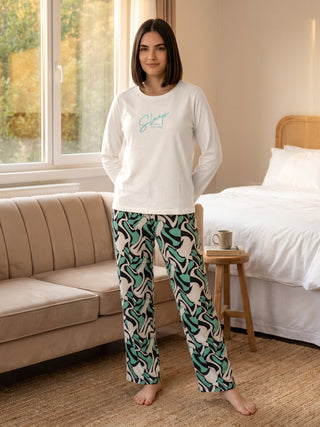 A woman stands in a bedroom wearing a white long-sleeved pajama top with "Sleep solves everything" printed on it and black, white, and green wavy-print pajama pants.
