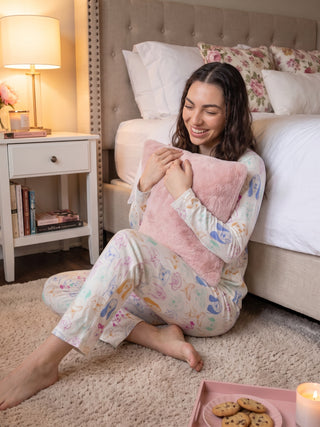 A woman sits on the floor in a cozy bedroom, wearing white pajama pants printed with colorful dog faces and holding a pink faux fur pillow.