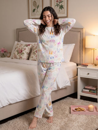 A woman in a white pajama set with a colorful dog print smiles and stretches in bed.