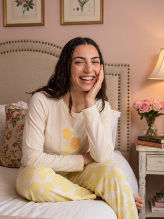 A woman poses on a bed wearing a cream long-sleeved sleep shirt with yellow flower print and matching cream pants with yellow flower print.