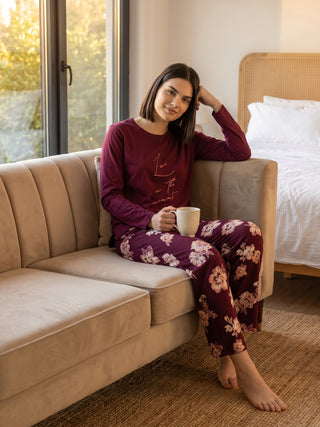 A woman in maroon floral print pajamas sits on a sofa, holding a mug.