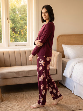 A woman stands in burgundy floral print pajama pants and a matching long-sleeved top, arms crossed.
