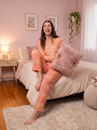 A woman is posing on a bed, wearing coral pajamas with a plaid print and a fluffy pink pillow.