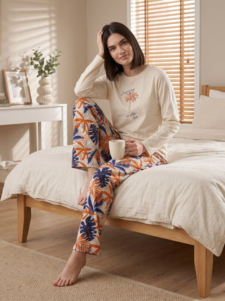 A woman in cream long-sleeve pajamas with a tropical leaf print poses sitting on a bed.