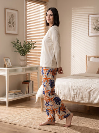 Women wearing a cream long-sleeved top and patterned pajama pants with an orange, blue, and white tropical leaf print, standing on a rug next to a bed.