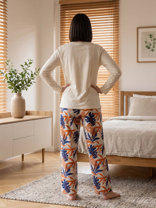 A woman stands facing away from the camera, wearing a cream-colored long-sleeved top and printed pajama pants featuring a tropical leaf motif in blue and orange.