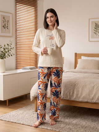 Women in a cream long-sleeve pajama set with a palm tree graphic, paired with orange and blue leaf-print pants, holding a mug.