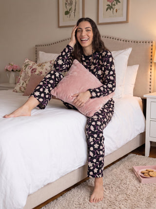 A woman sits on a bed in black pajamas with a floral print, holding a pink pillow.