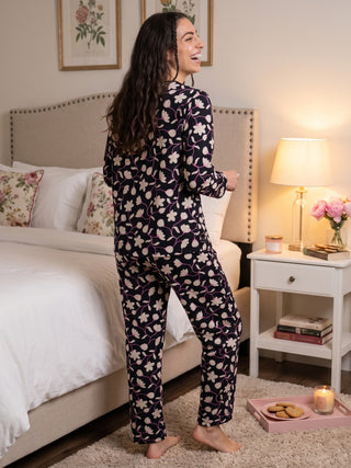 A woman in a black pajama set with a floral print stands in a bedroom.