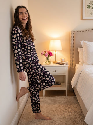 A woman leans against a wall, smiling, wearing a navy blue pajama set with a white and pink floral print.