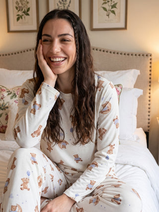 A woman sits on a bed wearing cream-colored pajamas with a teddy bear print.