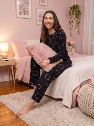 A woman in black pajamas with a white circle print sits on a bed, holding a pink pillow.