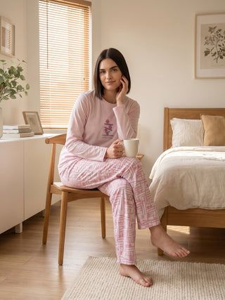 A woman in a pink pajama set with a geometric print sits on a wooden chair, holding a mug.