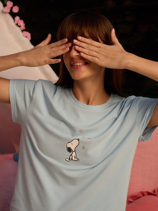 A woman in a light blue Snoopy pajama top, covering her eyes with her hands and smiling.