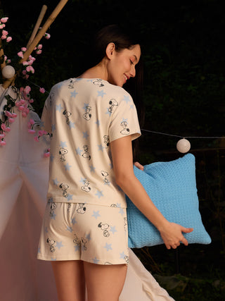 A woman in a cream-colored Snoopy-print pajama set consisting of a t-shirt and shorts, holding a blue cushion.