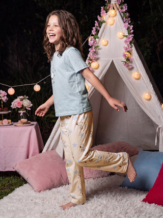 Girls wearing light blue Snoopy-print pajamas, standing on a fluffy rug in front of a decorated teepee, with a pink table laden with treats in the background.