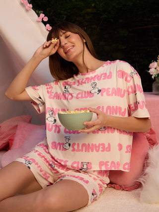 A woman in light pink Peanuts Club printed sleepwear, including a t-shirt and shorts, sits cross-legged holding a bowl of popcorn.