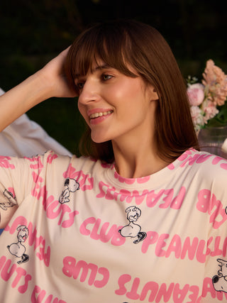 A woman wears a cream-colored, long-sleeved sleep shirt with a repeating pink "Peanuts Club" print and Snoopy graphics.