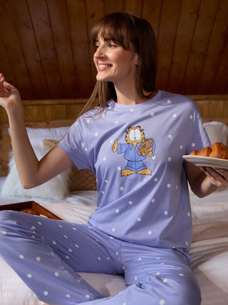 A woman in light purple polka dot pajamas with a Garfield print poses on a bed.