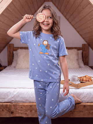 A young girl poses cross-legged on a rug wearing blue Garfield polka dot pajamas, covering one eye with a star-shaped cookie.