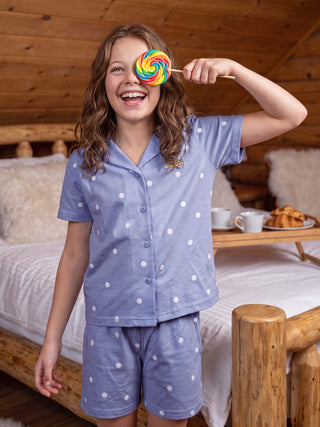 Girls in light blue polka dot pajamas, holding a rainbow lollipop over one eye while sitting on a bed.