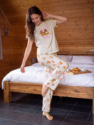 A young woman in cream-colored pajamas with a Garfield print smiles while striking a playful pose on a bed.